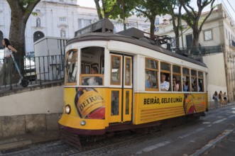 A yellow tram with Beirão advertising runs through a narrow city street, people on board, tram,