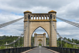 Historic bridge with distinctive arches and cables, surrounded by green landscape and a cloudy sky,