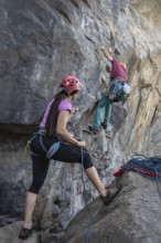 A woman stands at the base of a mountain, holding a climbing rope as her partner ascends to the top