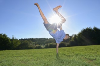 A boy doing a one-handed handstand on a grassy field with the sun shining brightly in the clear