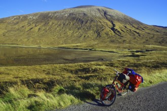 A loaded bicycle stands at the roadside in front of an impressive mountain landscape under a clear