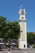 A tower clock with the Greek flag surrounded by trees and people in a relaxed atmosphere, Xanthi,