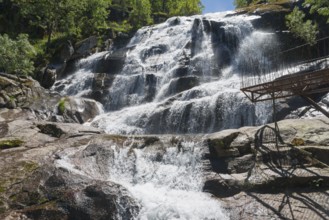 A large waterfall flows over rocks with a parapet in the foreground, surrounded by trees on a clear