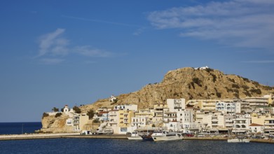 Coastal town with white houses and boats in the harbour, surrounded by hills under a clear blue