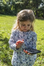 A little girl holds a smartphone and stands pensively on a sunny meadow