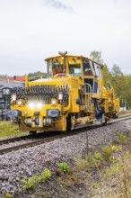 Yellow railway vehicle on tracks in an environment with industrial buildings, tamping machine,