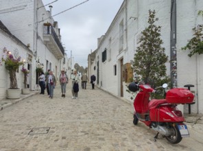 A picturesque street with white buildings, a red Vespa and people walking along, Alberobello,