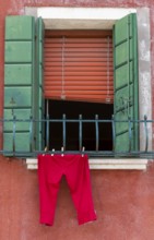 Half open window with green shutters and red trousers hanging in front of it, Burano, Venice,