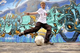 A ten-year-old boy plays with his football in front of a graffiti wall, football pitch in Germany