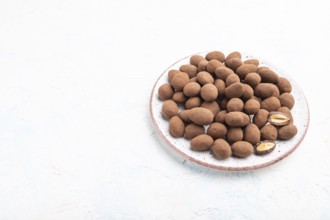 Almond in chocolate dragees on ceramic plate on white concrete background. Side view, copy space,