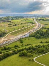 View of a large construction site next to a road through green fields and under a cloudy sky, A8