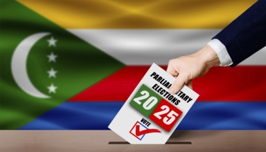 Hand holding ballot in voting ballot box with Comoran flag in background. Comoran parliamentary