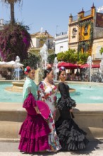 Three woman in colourful flamenco dresses, with flowers in their hair, stand in front of a fountain