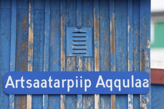 A blue sign inscribed with a Greenlandic street name on a weather-beaten wooden wall, remote Arctic