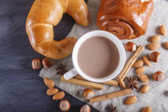 A cup of hot chocolate with nuts, buns and spices on linen napkin and black wooden background.