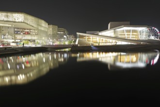 Modern buildings and opera house with glowing facades reflected in calm waters at night, Oslo,