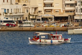 A small boat sails along the shore in a harbour with various buildings and boats, fishing boat,