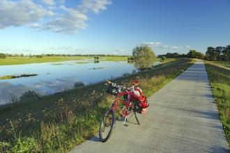 A bicycle stands on a concrete path next to a calm river under a clear blue sky with scattered