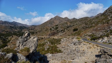 A road winds through a mountainous area with striking rock formations under a blue sky,