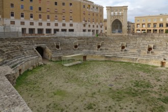 An ancient Roman amphitheatre made of stone with grass inside, surrounded by historic buildings,