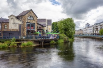 Historic buildings and cafés along a river with green vegetation and clouds, Sligo