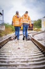 Two men in orange walking hand in hand on railway tracks in an unclear direction, track