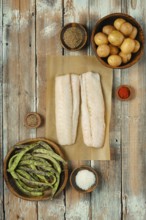 Two fresh cod fish fillets on parchment paper surrounded by various spices, green beans, and small