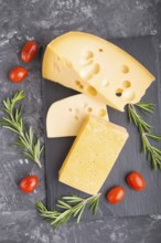 Various types of cheese with rosemary and tomatoes on black slate board on a black concrete