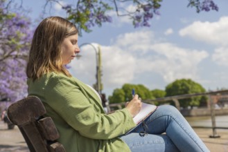 A young woman sitting on a bench in a park, writing in a notebook with a pen. The background