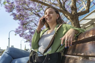 Happy woman with a green jacket enjoying a phone call outdoors, sitting on a bench under blooming