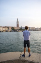 Young man in striped shirt and shorts on the banks of the Grand Canal, behind Campanile, Venice,