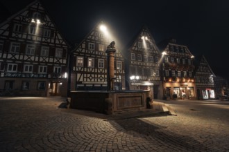 Illuminated square with fountain and half-timbered houses at night, cobblestones emphasise the