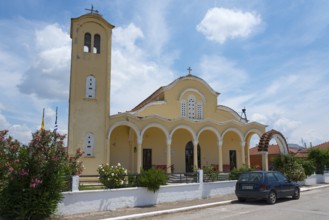 Church with yellow facade and bell tower under a blue sky, surrounded by flowers and a parked car,