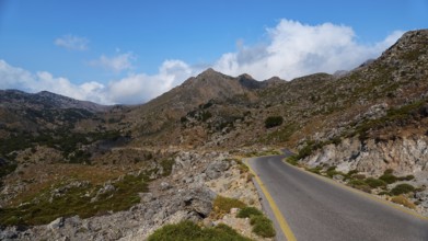 Asphalt road winds through a rocky mountain landscape under a blue, slightly cloudy sky,