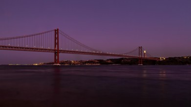 Twilight descends on a calm river with a suspension bridge standing majestically against a purple