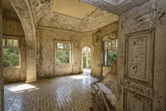 An abandoned room with decorated tiles, an arched entrance and open windows shows clear signs of