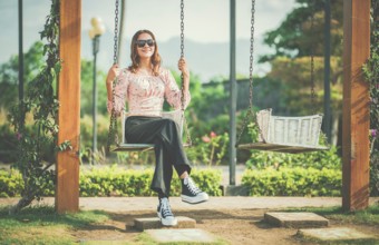 Cheerful girl in glasses relaxing on a swing in a natural park. Happy young woman sitting on a