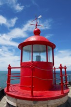 Red lighthouse with round roof on a coastal platform, blue sky and sea in the background, Fort São