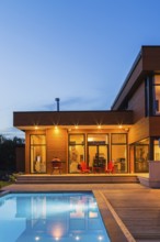 Modern cube style home with stained horizontal wood cladding and in-ground swimming pool at dusk,