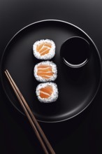 Three sushi rolls filled with fresh salmon are neatly arranged on a black plate. A small bowl of