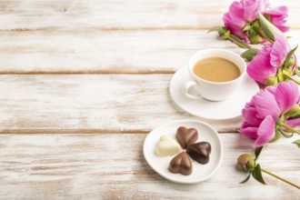 Cup of cioffee with chocolate candies, pink peony flowers on white wooden background. side view,