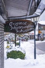 Company sign for Black Forest ham on a snowy street with shops, Enzklösterle, district of Calw,