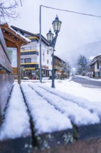 Snow-covered park bench in the centre of a wintry village with traditional houses and street lamps,