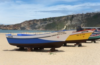 Several colourful fishing boats lying on the sandy beach of Nazaré with hills in the background,