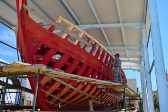 A partially built red wooden ship in a covered shipyard with a labourer working on it, Patmos