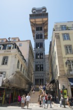 Tower-like lift between old buildings, people walking up the stairs, clear sky, sunny weather,