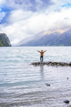 Person standing with outstretched arms on rocks by the lake with mountain backdrop under dramatic