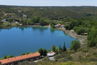 View of a tranquil lake with surrounding houses, trees and hills under a blue sky, view from the