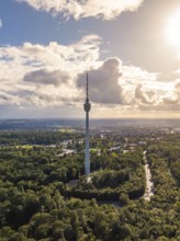 The television tower rises above a large wooded area under a partly cloudy sky in sunlight,