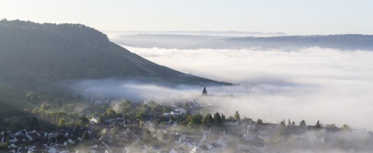 A village lies in the morning mist in front of a hill, with a church rising out of the mist,